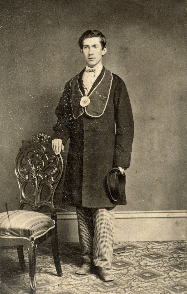 A young man poses in his fraternal sash with his initials embroidered on one side and a flower in the center