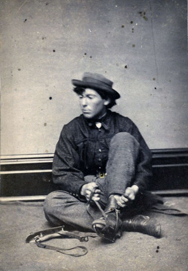 A young man sits on a blanket upon the floor of the photographer's studio, his left leg raised and hands pulling the leather straps that fasten an ice skate to his ankle-high boot