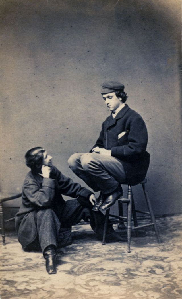 A young man wearing a cap sits on a stool, looking down at another young man seated on a cushion and returning his gaze