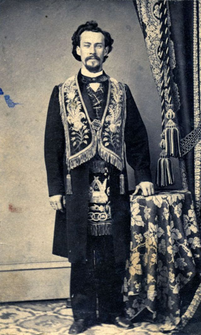 A bearded man stands before the camera operator wearing the embroidered accouterments that signal his membership in the Masons