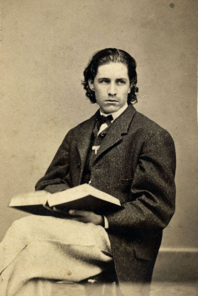 A young man with a ribbon pinned to the lapel of his vest sits in a chair and holds a book