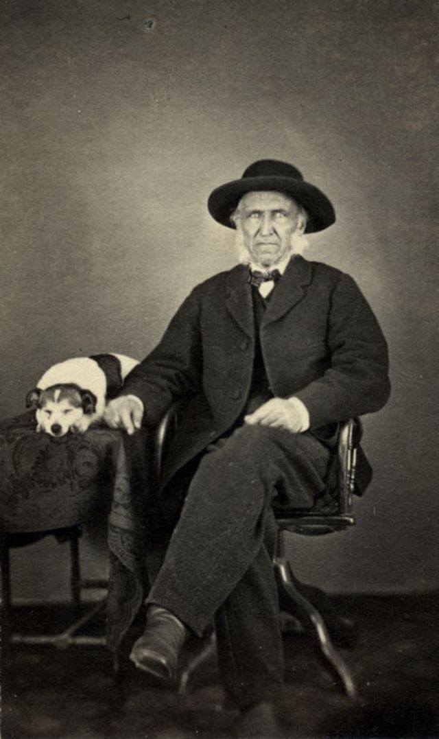 An elderly man sits in a chair with his right arm resting on a cloth-covered table on which is seated a dog. The dog is asleep, head resting on paws