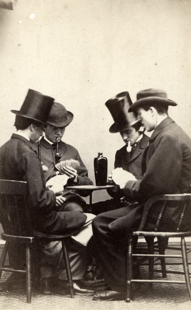 Four gentlemen with pipes clenched in their teeth sit with playing cards and a bottle of gin