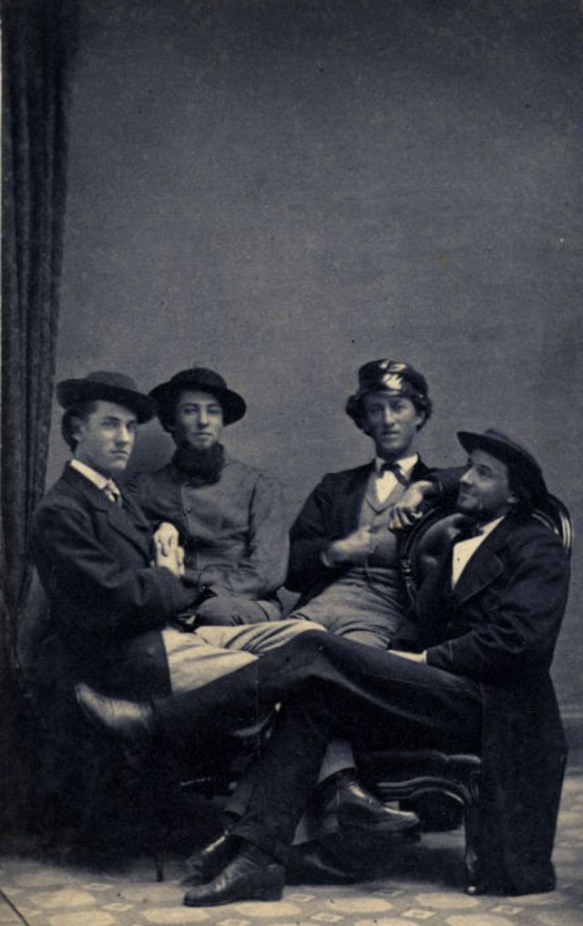 Four young man, all with smiles or hints of smiles upon their faces, relax in chairs and a bench artfully arranged in the photographer's studio