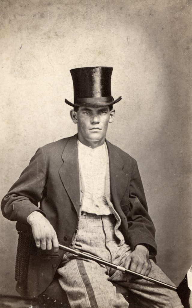 A clean-shaven young man wearing a white collarless shirt, vest, striped checked pants and coat sits with a stick and top hat