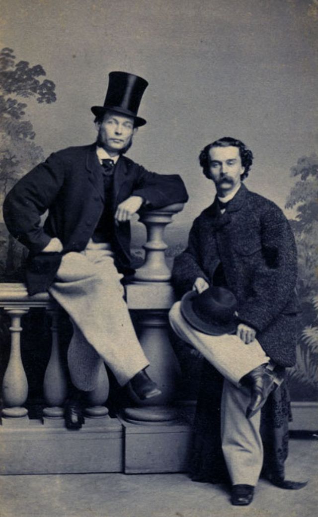 Two gentlemen sporting beaver skin hats strike jaunty poses as they sit on a studio balcony prop