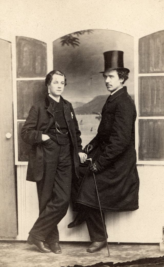 Two well-dressed men pose in front of a painted background featuring an open window