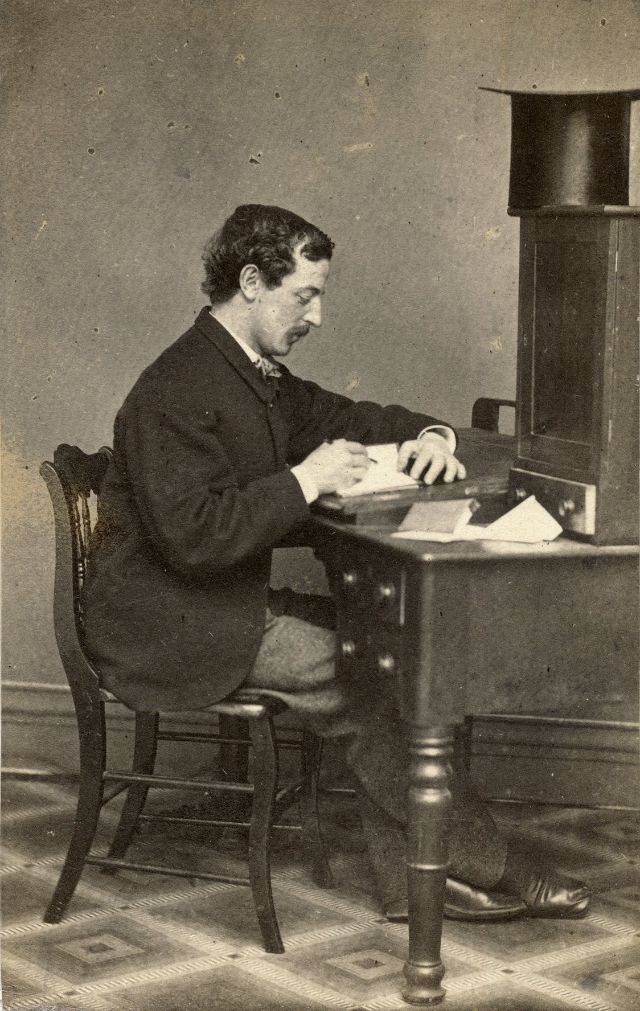 A man sits at a desk and writes in a small volume similar in size and thickness to a journal. His top hat rests on top of the cabinet mounted to the desktop