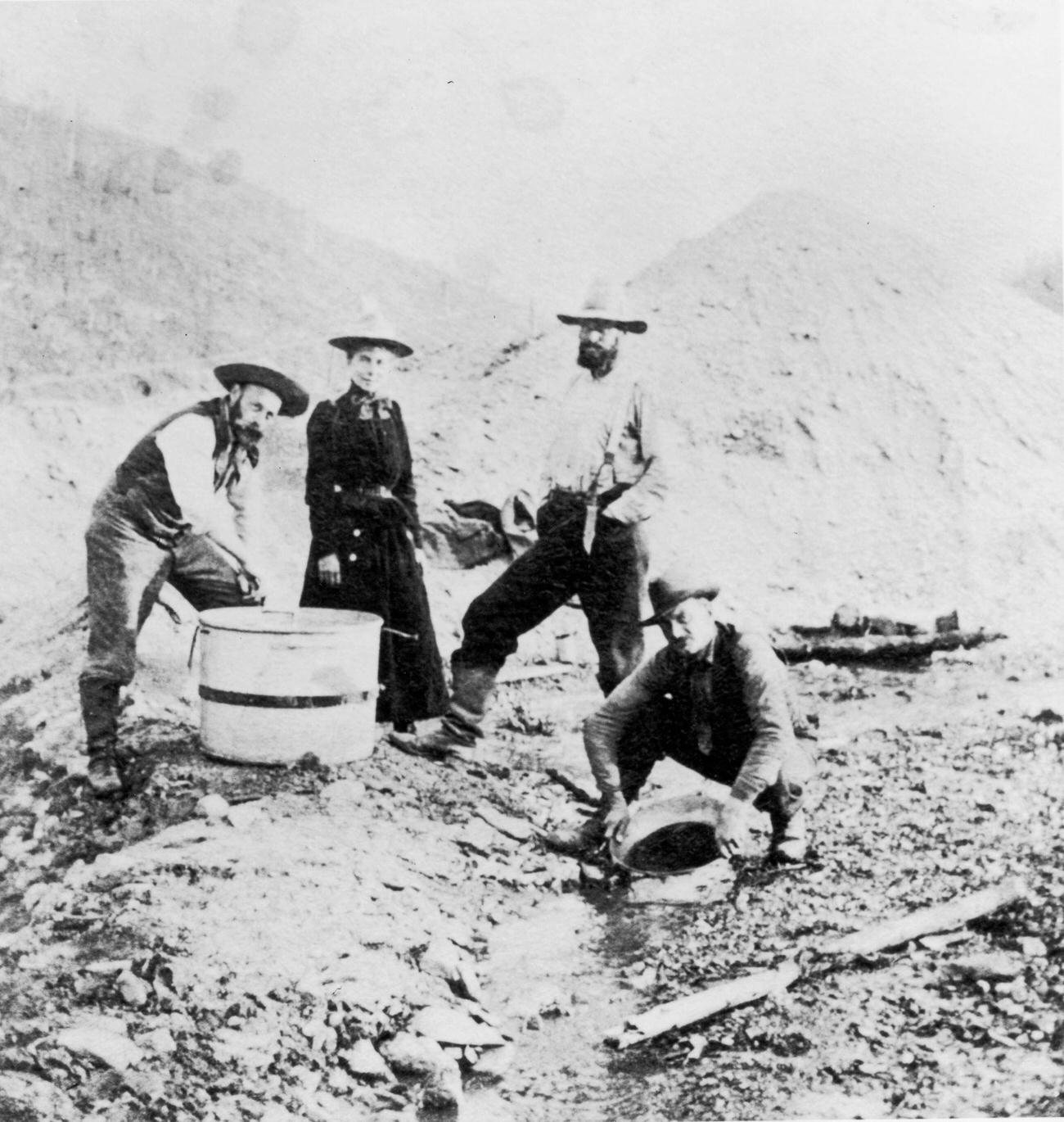 Three men and a woman panning for gold in a riverbed during the Klondike Gold Rush, Alaska.