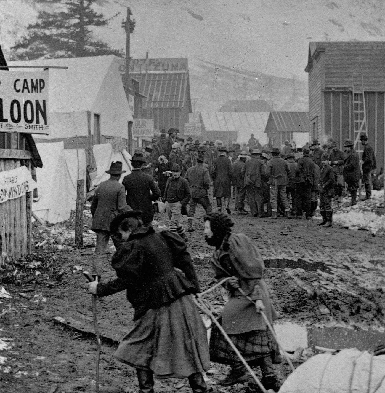 Two female prospectors pulling a sled through a Klondike gold fields boomtown.