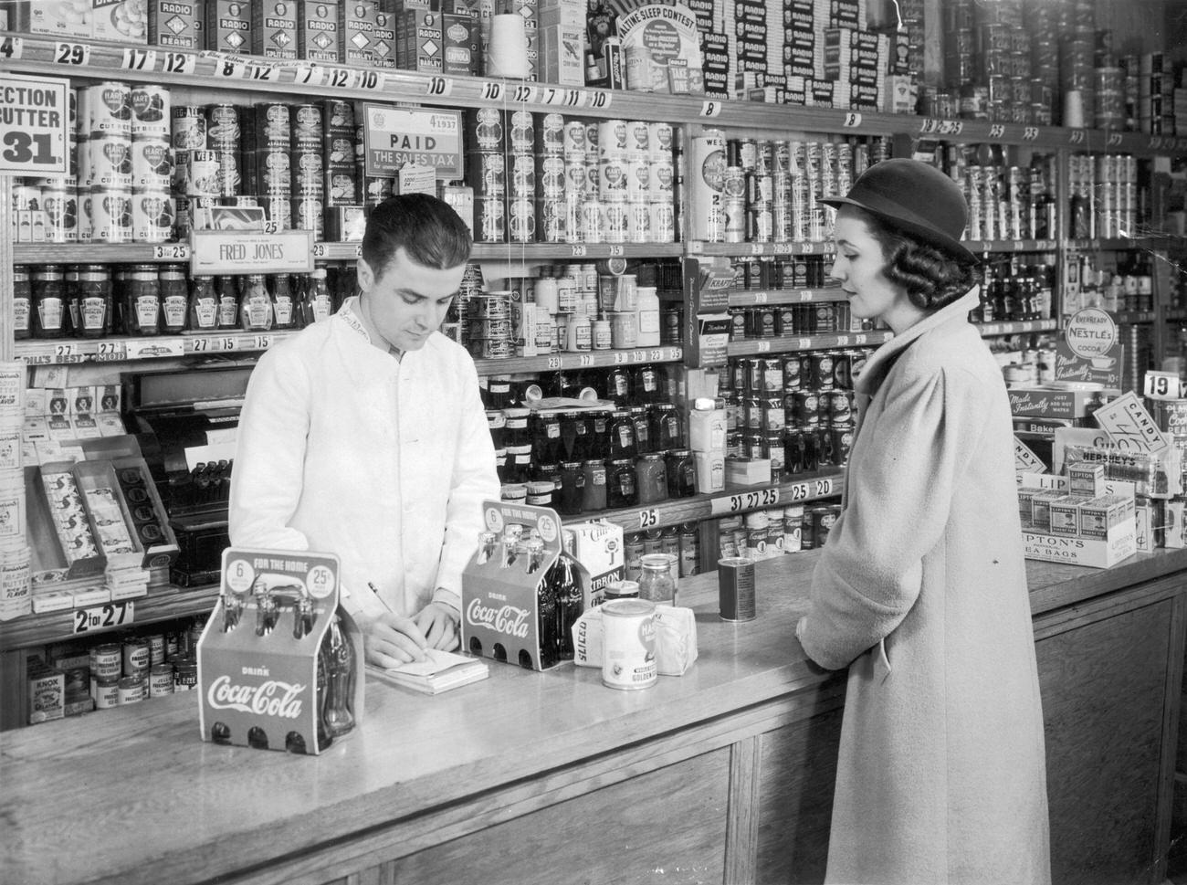 A housewife at a grocery counter with two six-packs of Coca-Cola, bread, and canned goods, circa 1935.