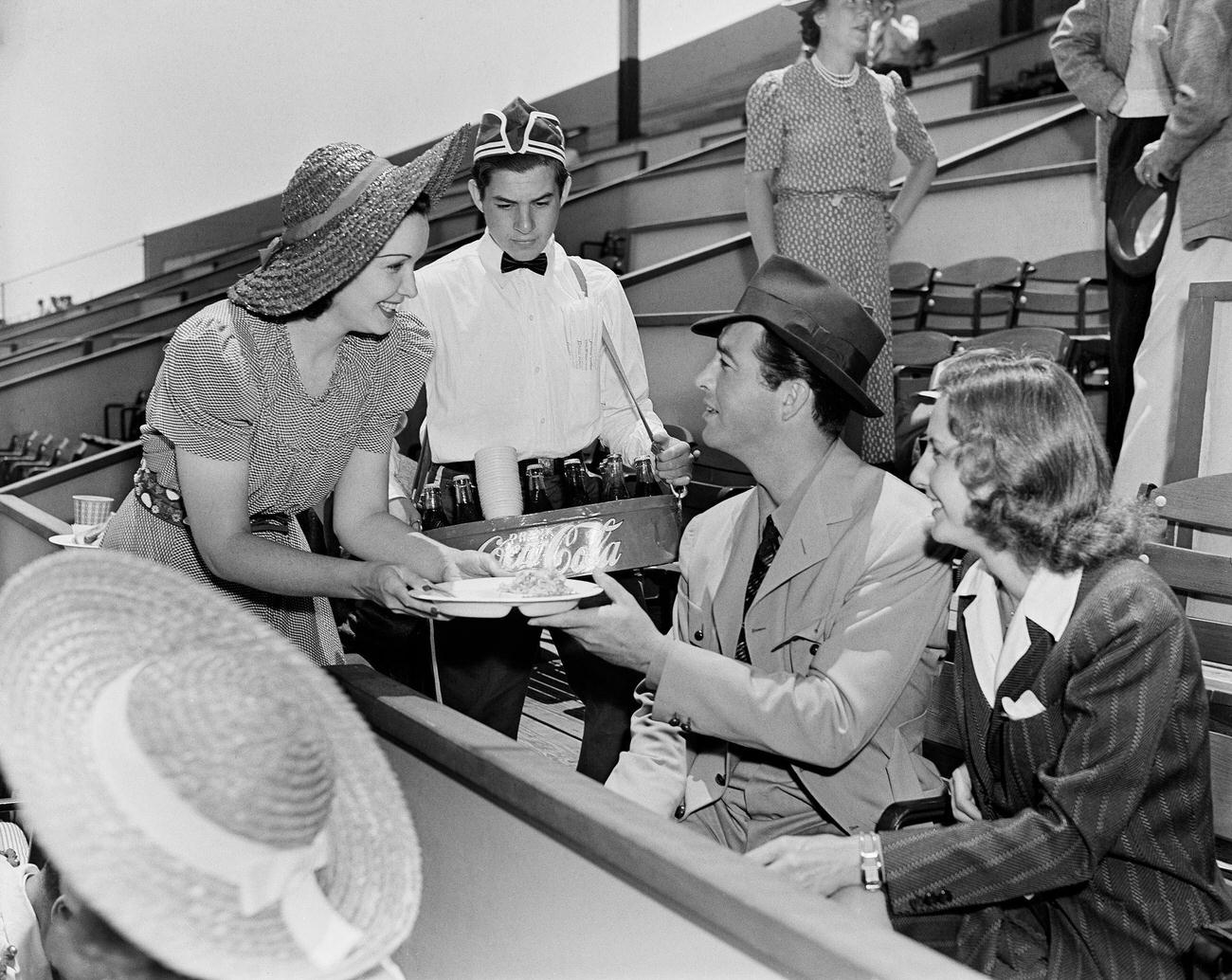 Actors Robert Taylor and Barbara Stanwyck offered food and Coca-Cola at a game in Los Angeles, circa 1940.