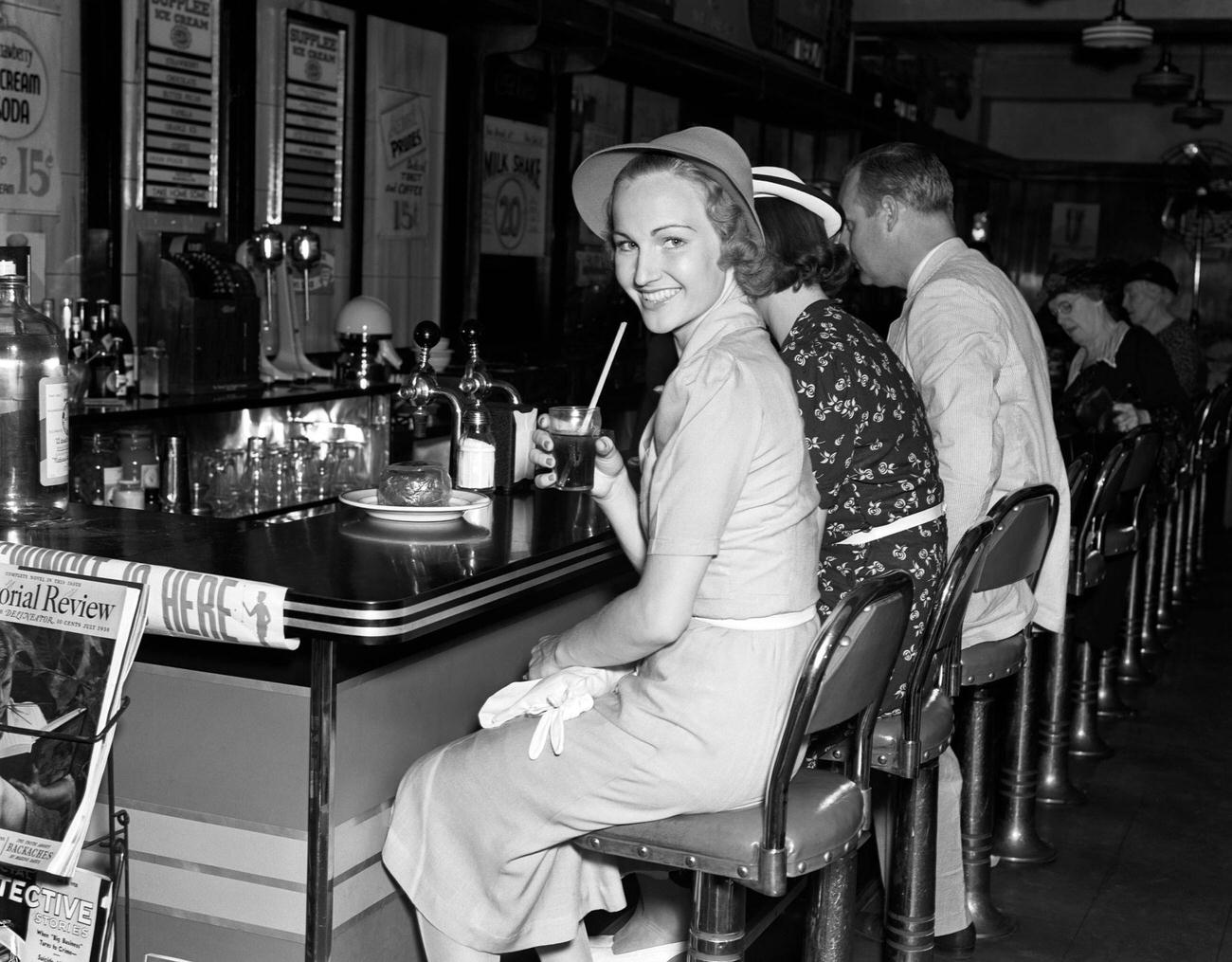 Smiling women in linen dresses with a glass of cola at a soda fountain, 1940s.