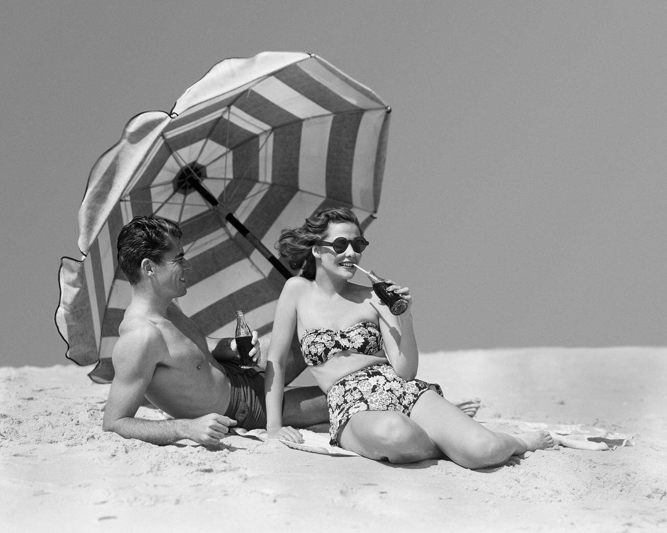 Couple sunbathing and drinking soda by a beach umbrella, 1950s.