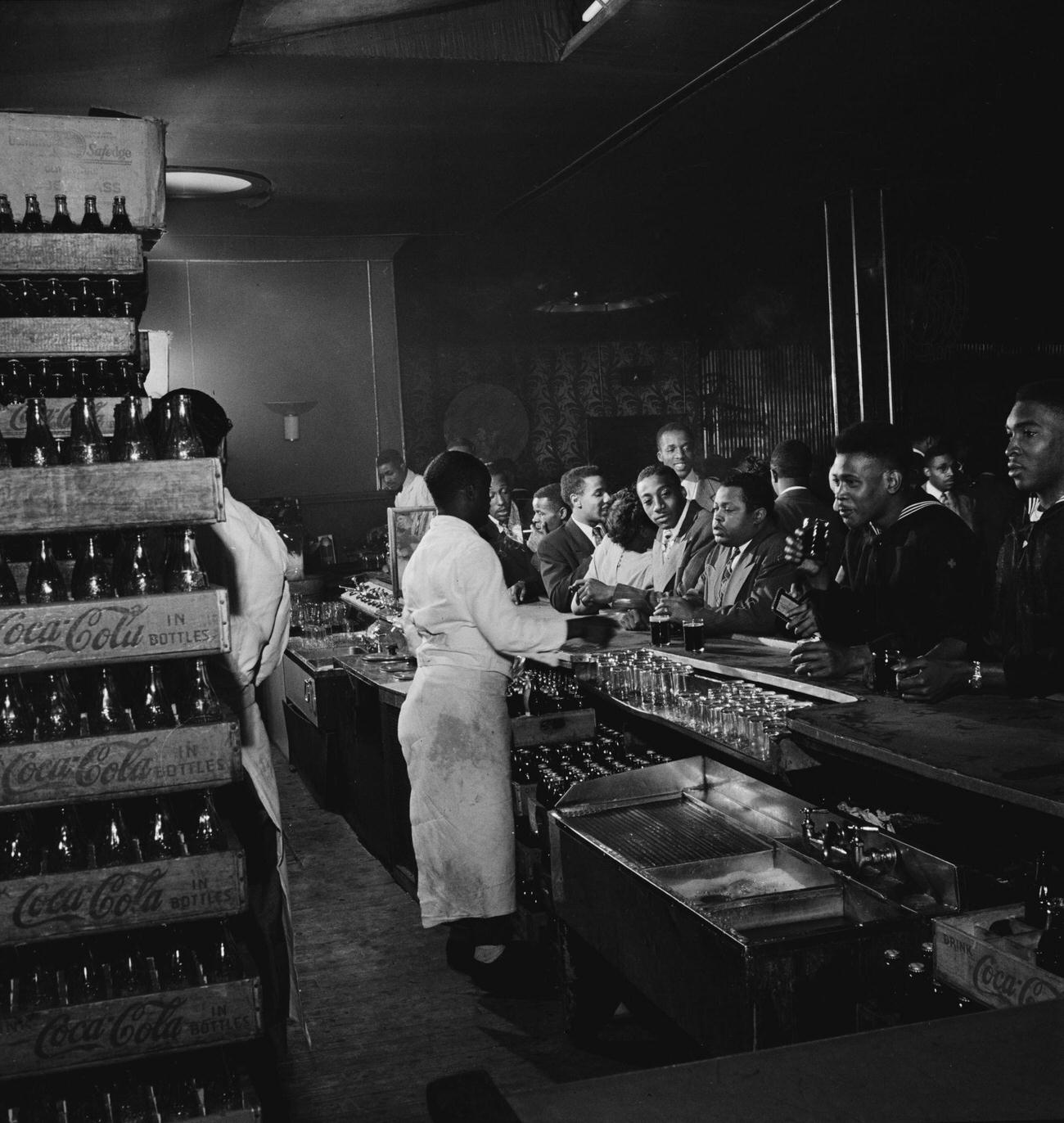 Coca-Cola crates stacked behind a bar with white-coated bartenders, location unspecified, circa 1950.