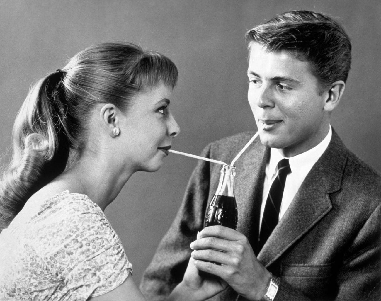 Teenage couple sharing a single bottle of soda with two straws, 1950s.