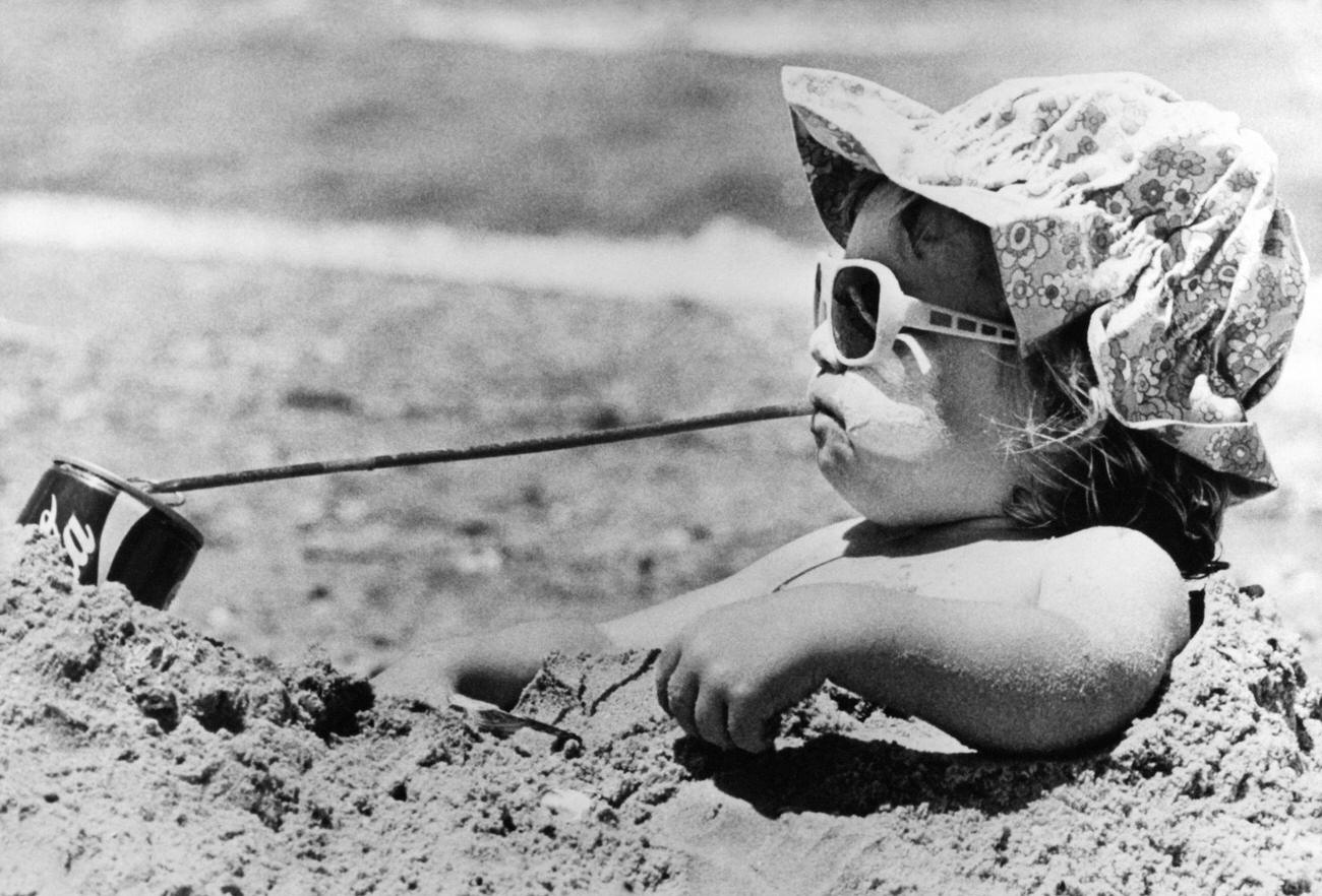 Young girl buried in beach sand drinking a cold beverage, Australia, circa 1960.