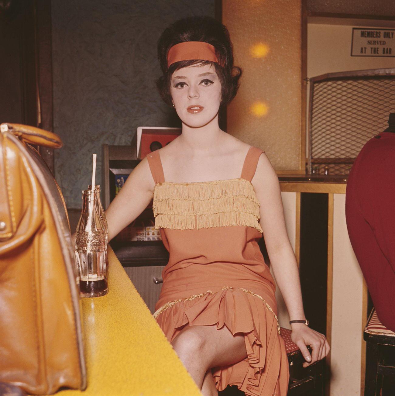 A young girl in an orange dress drinking Coca-Cola at Whisky A Go-Go Club in London, 1962.
