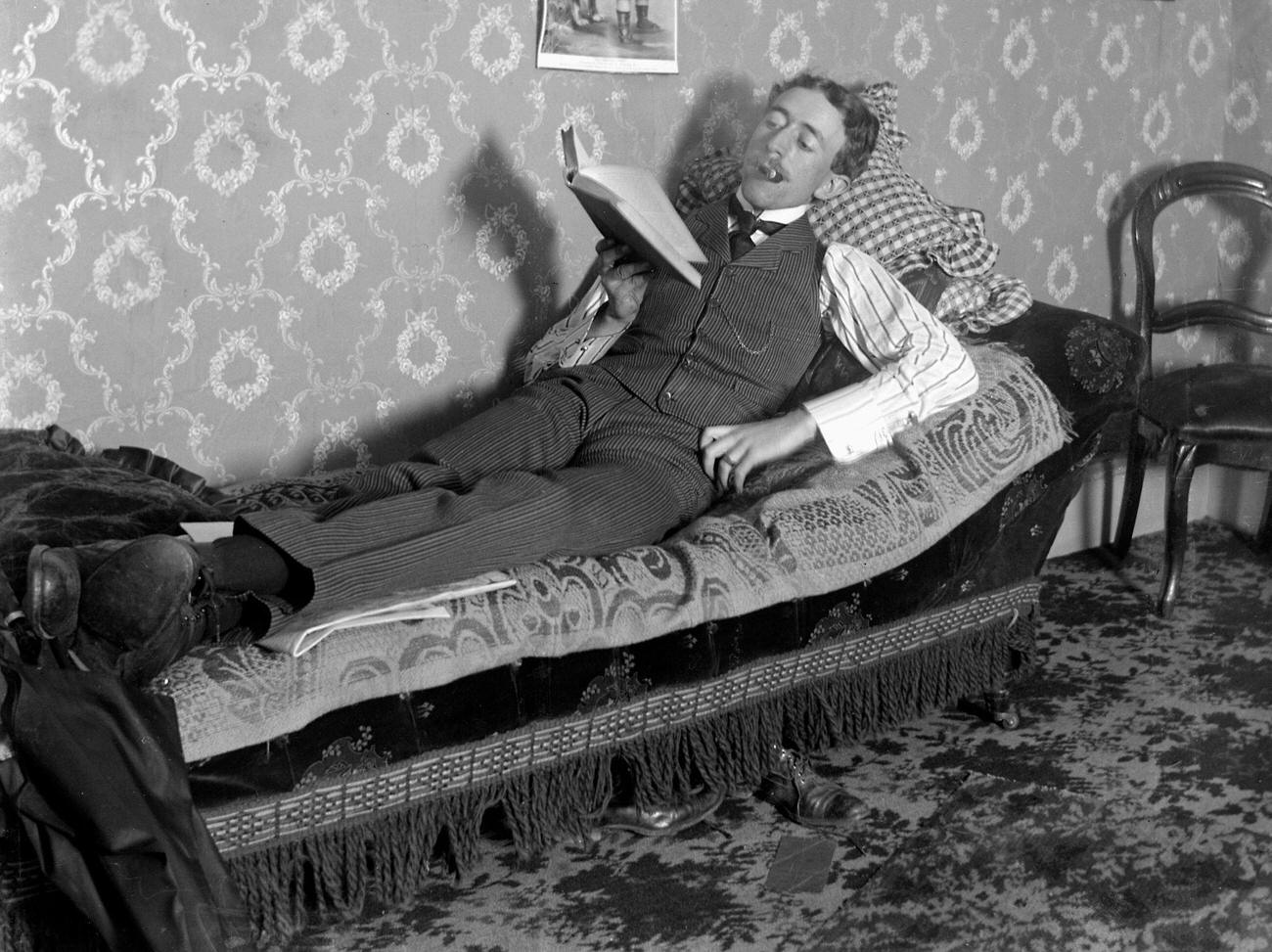 A man reading and smoking on a fainting couch, circa 1900.