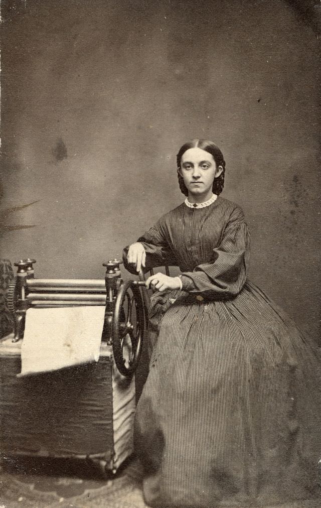 A young woman turns a wheel connected to a set of rollers. The sheet of paper suggests the hand-operated machine was used in some aspect of paper production or printing