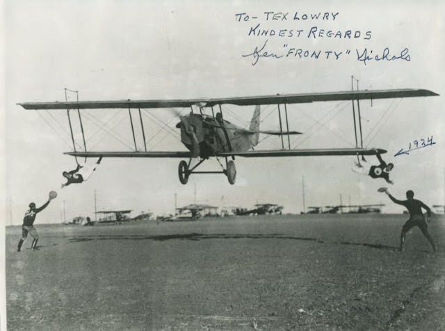 Two Black Cats hanging from the lower wing skids are about to snatch up objects from the two people on the ground. Inscription reads, “To Tex Lowry, Kindest Regards, Ken ‘Fronty’ Nichols.”