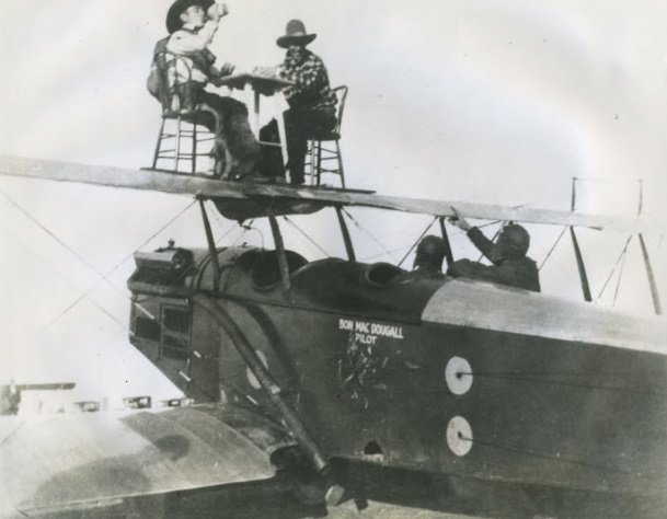 Bon MacDougall pilots the plane with Jack Frye pointing to “cowboys” Al Johnson (drinking from a bottle) and Ken “Fronty” Nichols who seem to be playing a friendly game of cards on the upper wing.