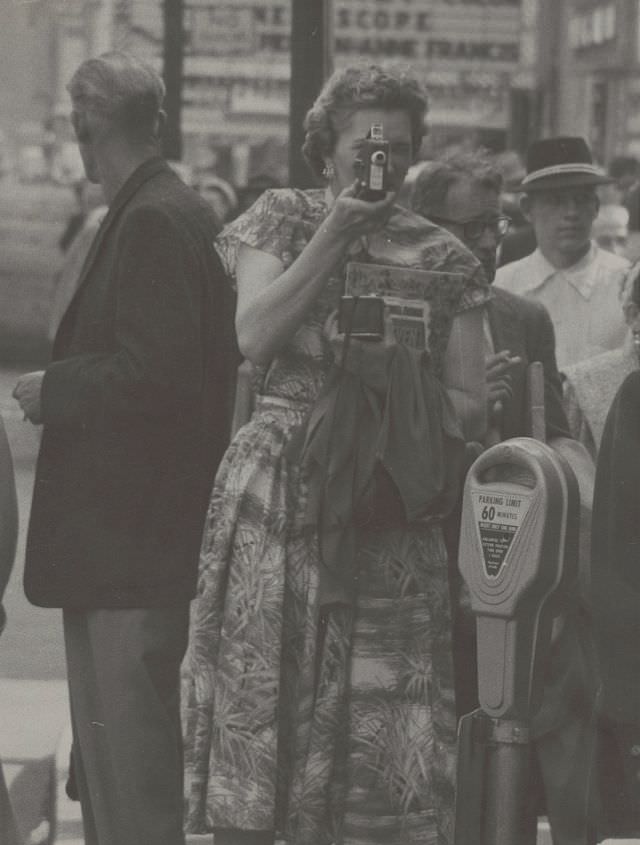 Woman holding a home movie camera while walking on New York City sidewalk, 1956