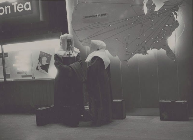 View of two nuns next to map of the United States with route markers and lights, suitcases on ground, November 1957