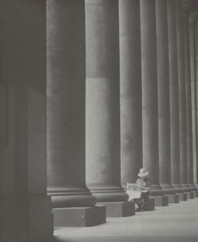 Woman reading newspaper while seated beside building columns, January 27, 1950