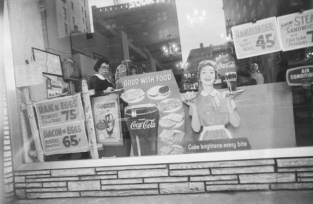 Woman at cash register viewed through diner show window with buildings reflected, February 1959