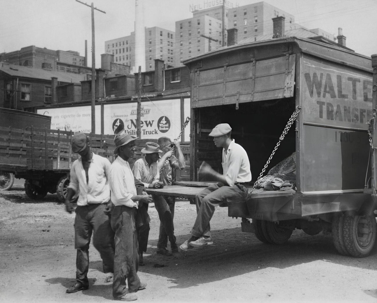 A group of men playing craps, one sitting on a truck's tailgate, at a transport facility on the banks of the Scioto River with the Neil House hotel in the background, Columbus, Ohio.