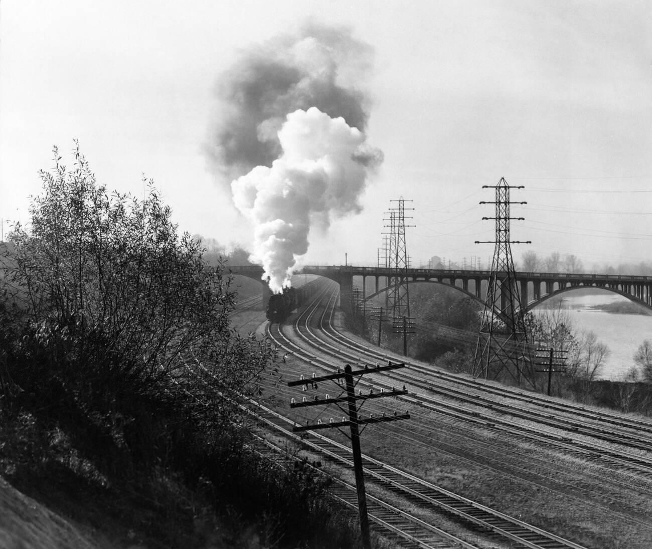 Aerial view of a train traveling along a river under a bridge, billowing smoke near Columbus, Ohio, in the 1940s