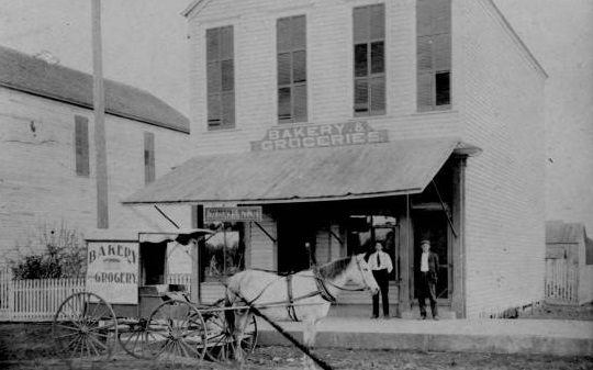 Bakery and Groceries building with two men posing, 1880s