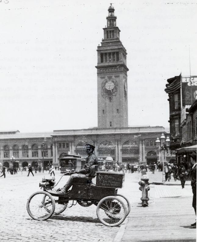 Lester L. Whitman at the tiller of a Curved Dash Olds Light Runabout in front of the Ferry Building, July 6, 1903