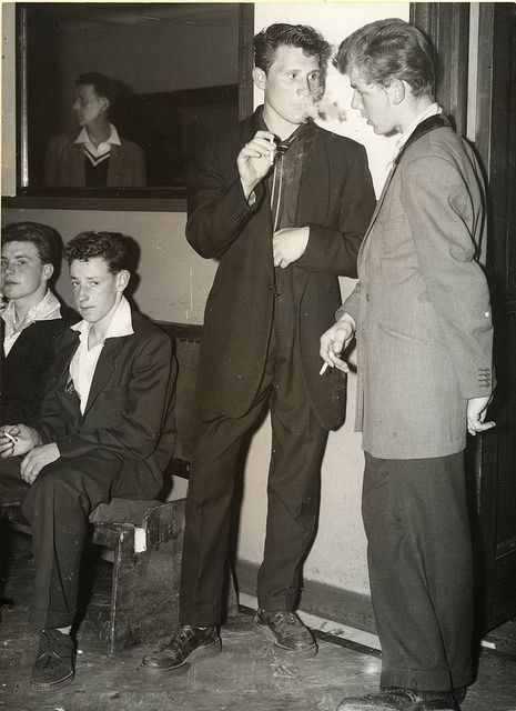 The Stag-Line at a Dance Hall. The Teddy Boy in the centre is wearing an Edwardian outfit, which may have cost him as much as £50.00 ( very expensive at the time). Note the Long Jacket with ‘Shawl’ collar and single link button fastening (nearly coming off), Drainpipe trousers, thick Crepe soled shoes (‘Creepers’) and parallel-striped ‘Slim Jim’ tie. The boy on the right has a ‘Tony Curtis’ haircut and is wearing crepe soled suede shoes. – July 1955.