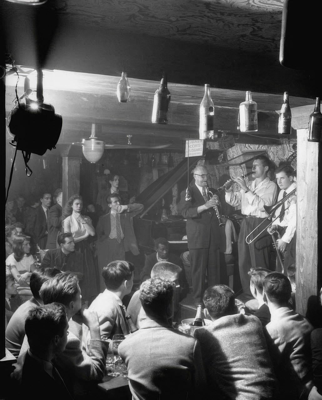 Jam session in shadowy cellar of Vieux Colombier attracts a crowd of Americans (foreground). Their compatriot, Clarinetist "Mezz" Mezzrow (left), is the big attraction. On such forays, the boys save money by dividing a bottle of champagne eight ways. In Paris, American teen-agers ignore Eckstine and Sinatra records as démodé, prefer French Crooners Charles Trenet and Yves Montand. They have a favorite French tune, Fou de Vous (Crazy About You).