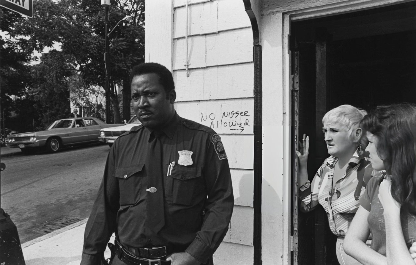 A police officer and two women in a doorway beside racist graffiti, Boston, 1975.