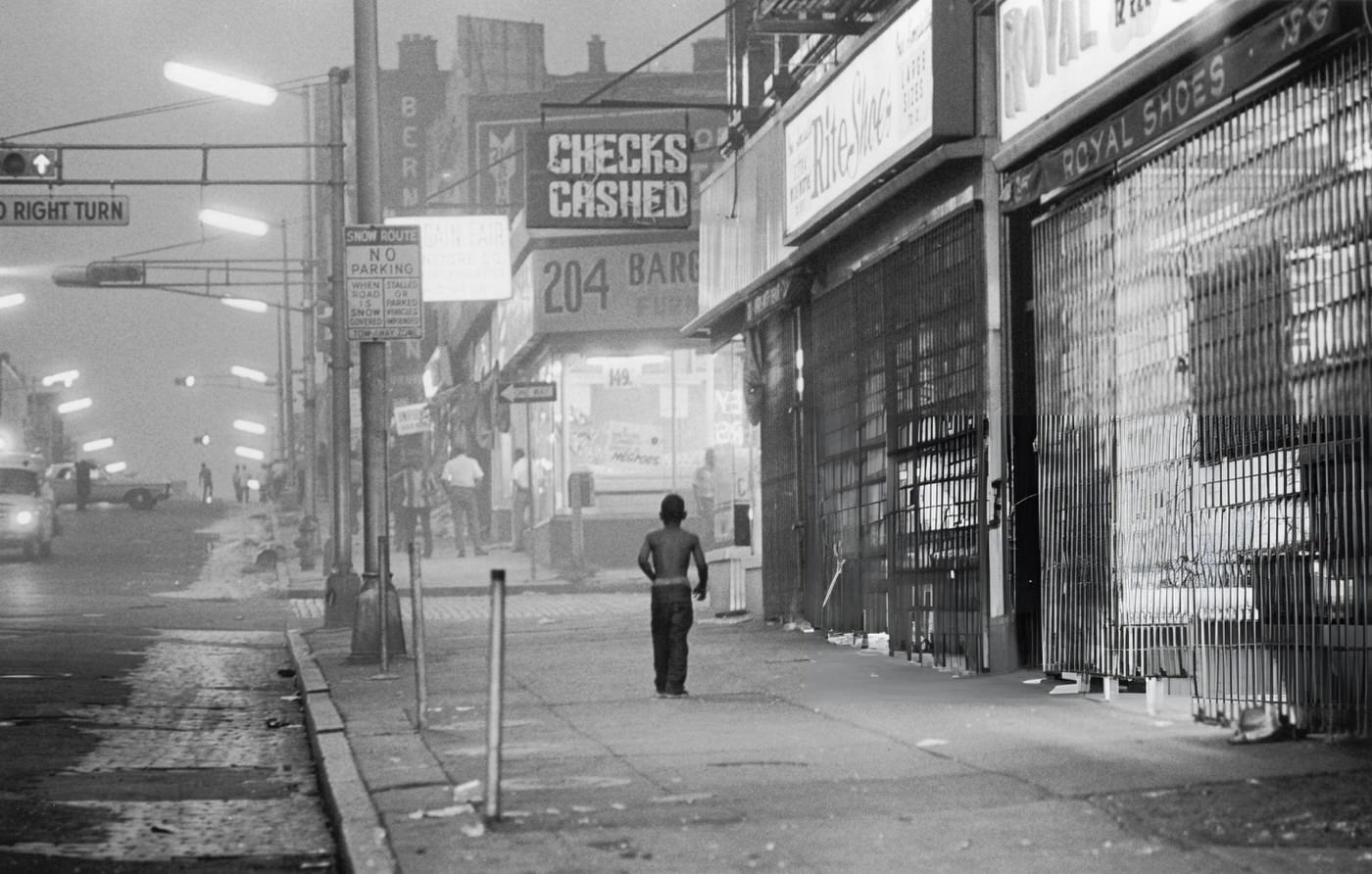 A young boy walks down the sidewalk past shuttered storefronts towards a furniture store
