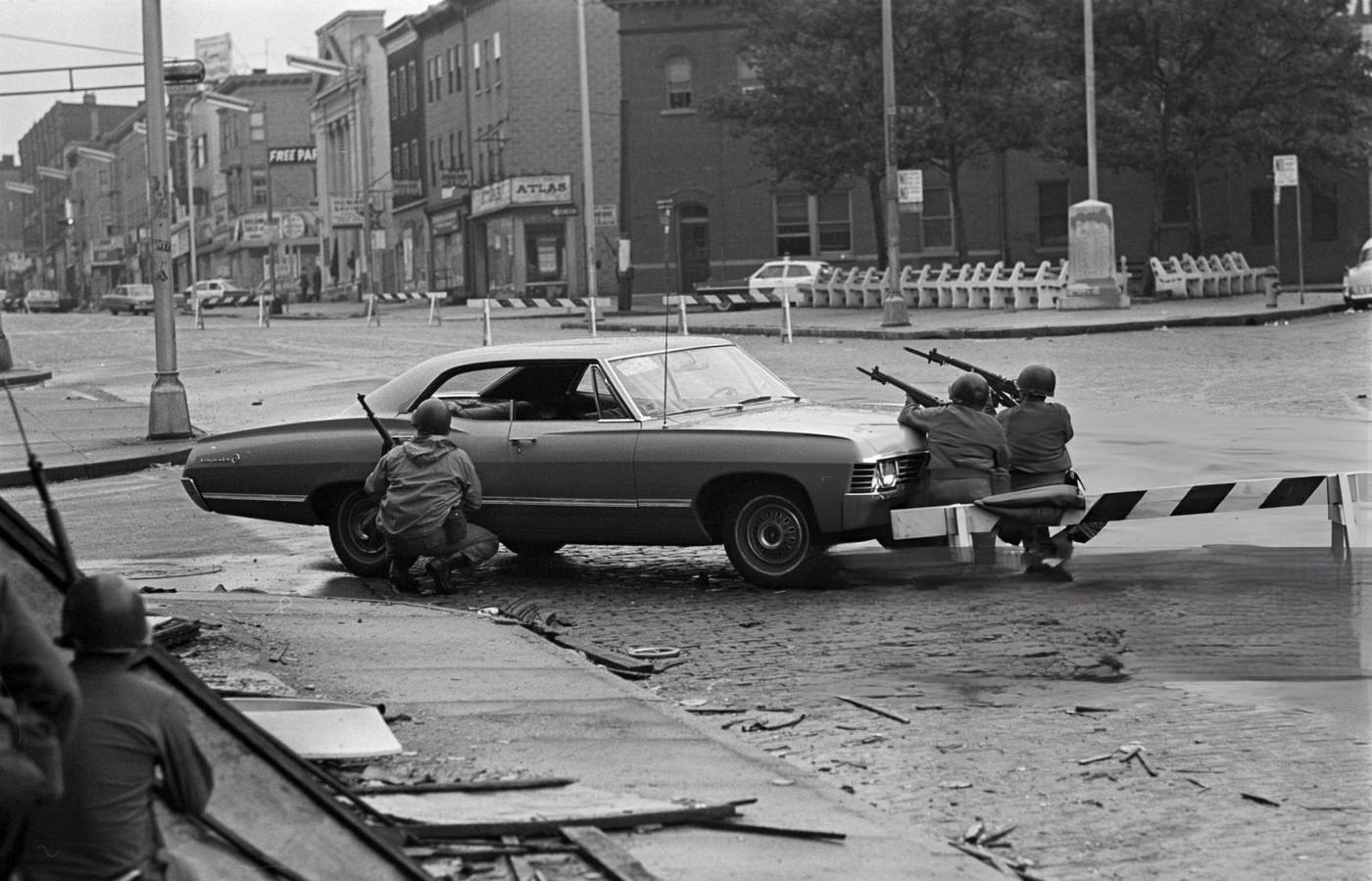 National Guard soldiers taking cover behind a car, guns pointed towards the street