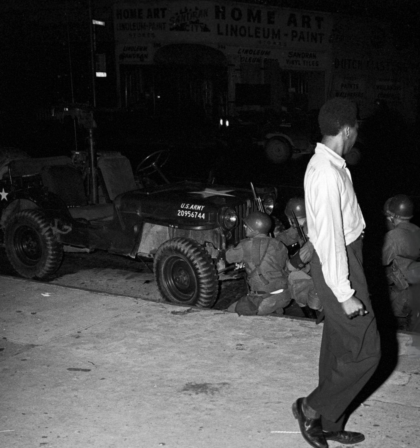 A young African American man walking on a sidewalk looks at a group of National Guard soldiers, 1960s.