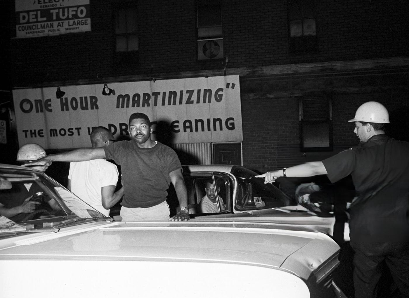 A National Police officer searches an African American man in front of a parked car