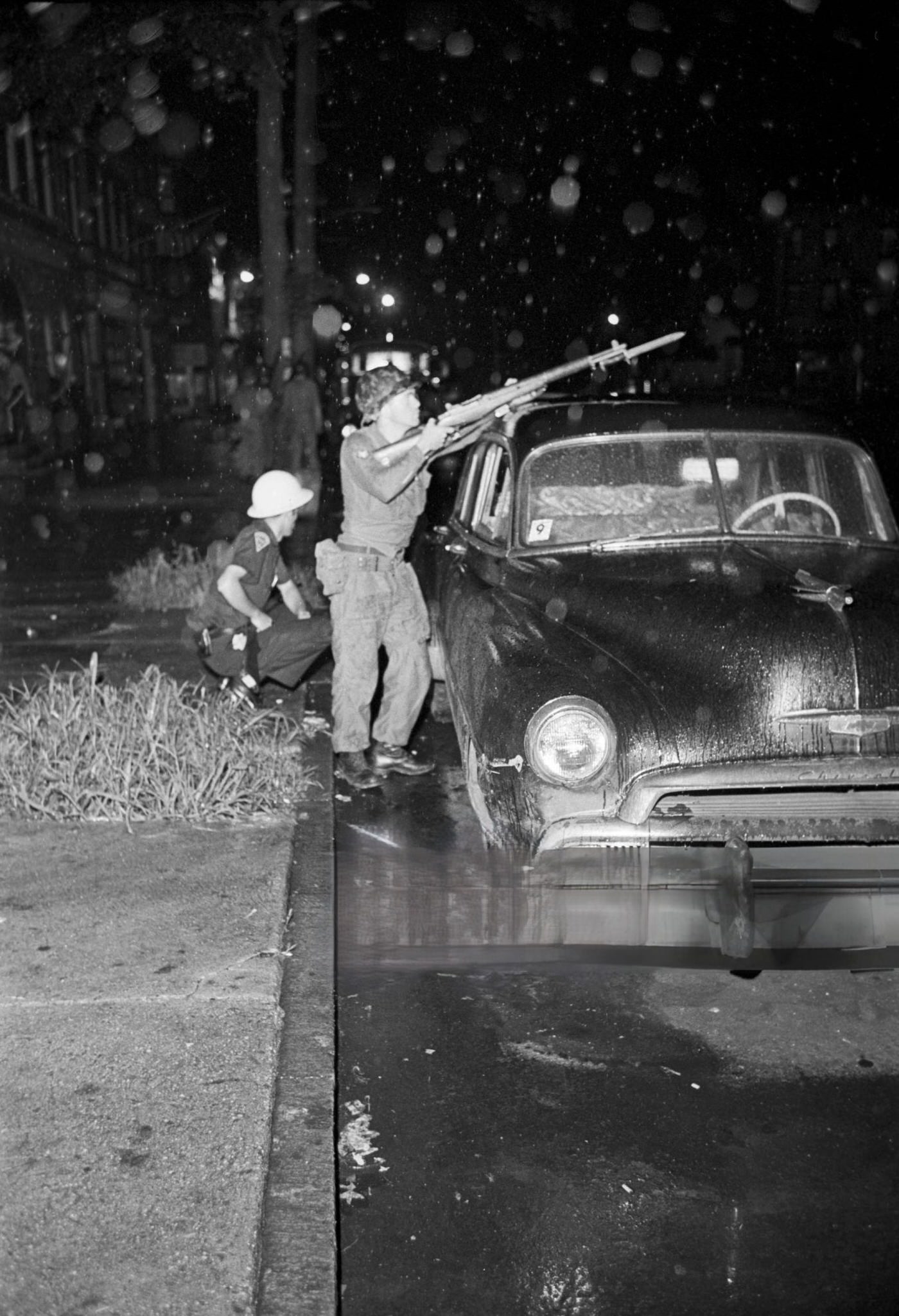 A National Guard soldier stands with a gun pointed towards the street next to a police officer crouched behind a jeep
