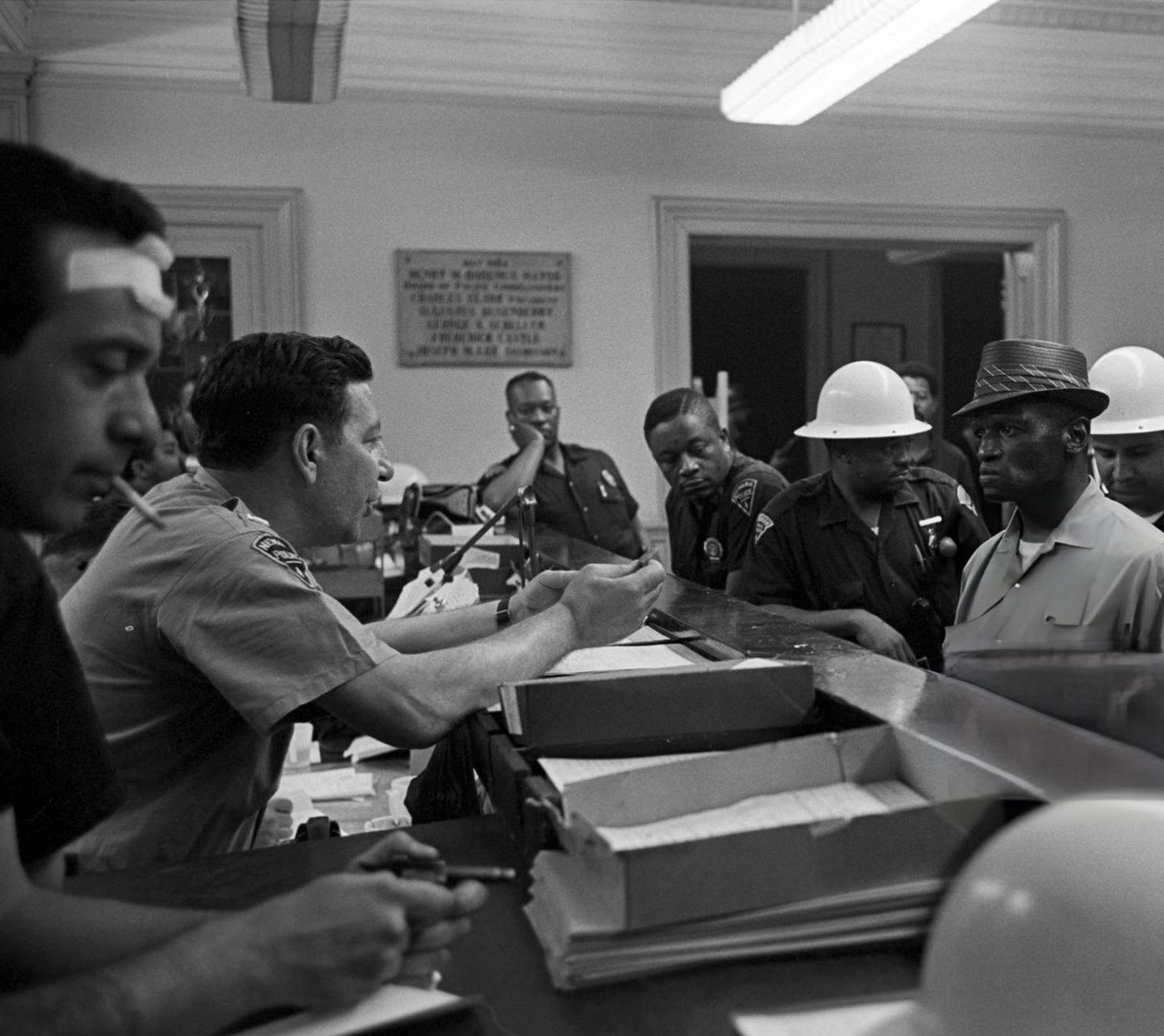 An African-American man in a hat under arrest, standing in front of a counter surrounded by Newark police officers, 1960s.