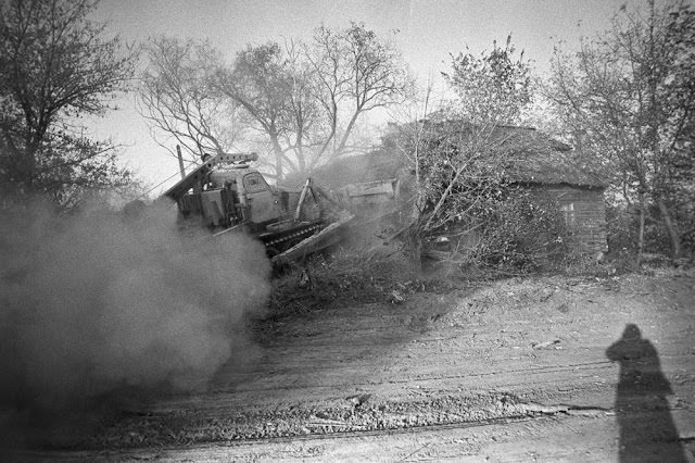 A bulldozer digs a large trench in front of a house before burying the building and covering it with earth.