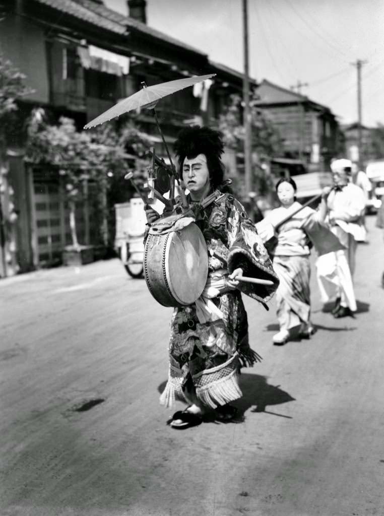 A Japanese man in costume beats the drum as he leads a musical parade of men and women who are walking advertisements featuring placards for restaurants through the streets of Tokyo, 1947.