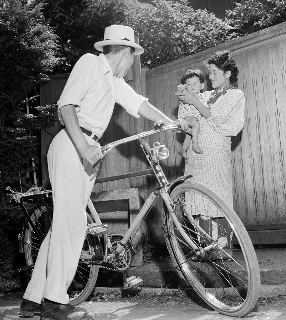 Baby Misuko, held by her mother Umeko, waves goodbye to papa-san as he goes to work on his bicycle, from their home in Tokyo, 1948.
