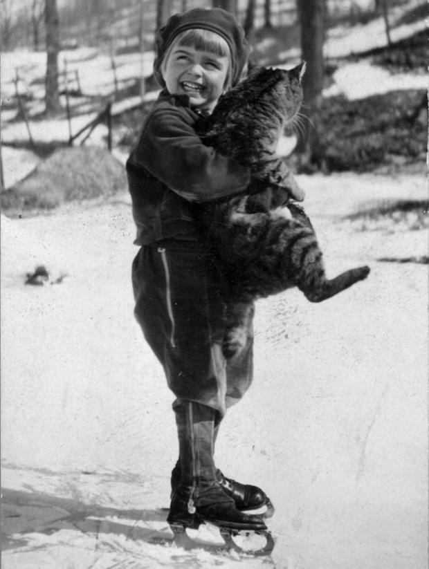 A young boy on skates clutches a large tabby cat, 1940.