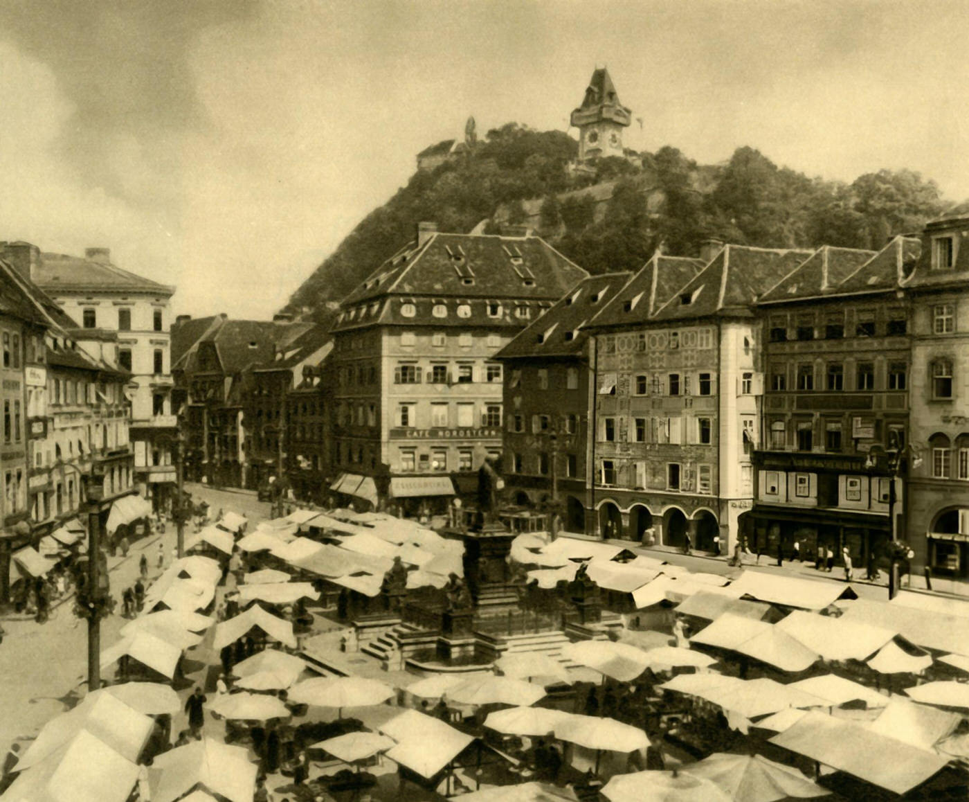 Market stalls in the center of Graz, with the clock tower of the Schlossberg beyond, Austria, 1935.
