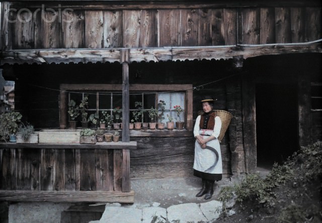 A woman standing outside a house with a scythe and basket, Heiligenblut, Austria, 1929.