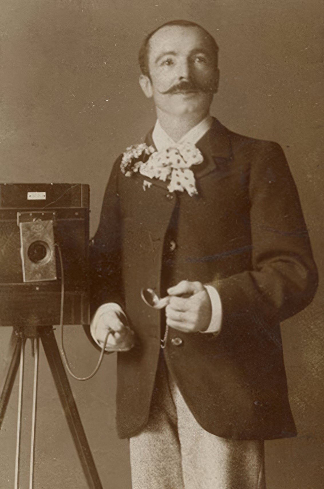 This gentleman has passed dapper and is approaching a lampoon representation of an artiste! The floppy polka dot bow tie and bunch of flowers might be his everyday dress but it feels to us as if he is having some fun or playing a part. He holds the bulb of a pneumatic shutter mounted on a large view camera. His pocket watch is in hand to time the exposure.
